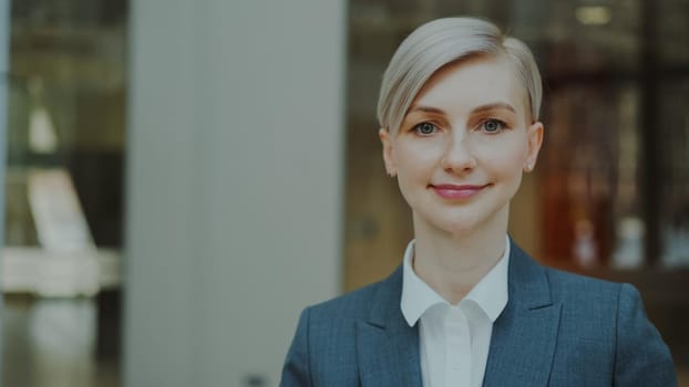 Close-up portrait of successful blonde businesswoman smiling and looking into camera in modern office indoors