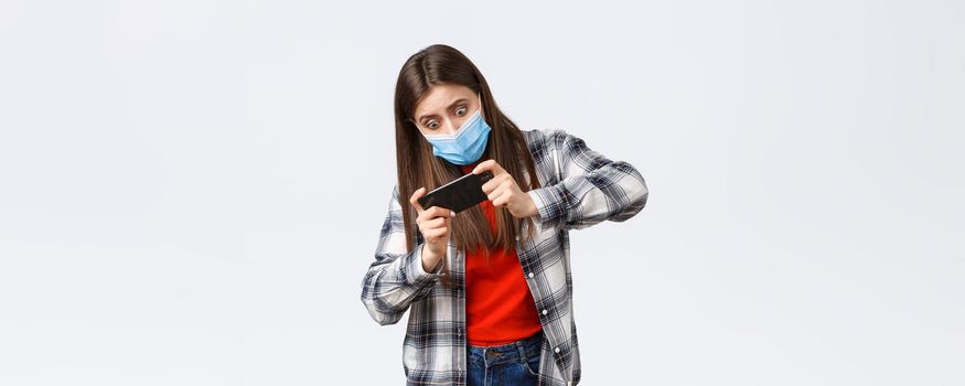 Different emotions, covid-19 pandemic, coronavirus self-quarantine and social distancing concept. Focused and entertained woman in medical mask playing mobile game, tilting body, difficult level.