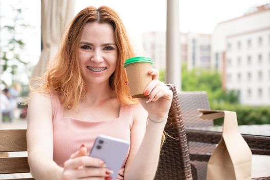 Young red-haired woman with braces on her teeth drinks coffee on a summer veranda