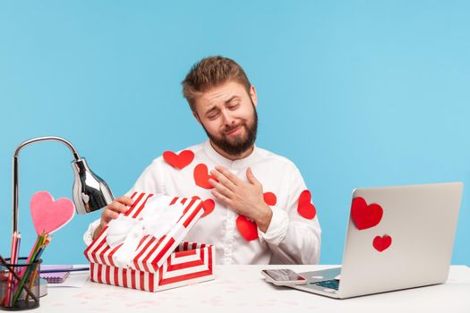 Cute pleased man with beard in white shirt with red sticky hearts unboxing present and looking inside with smile, satisfied with gift. Indoor studio shot isolated on blue background