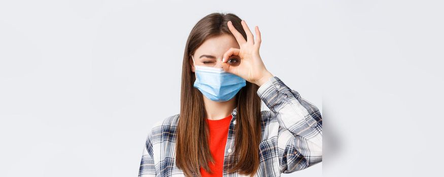 Coronavirus outbreak, leisure on quarantine, social distancing and emotions concept. Close-up of optimistic good-looking woman in medical mask show okay sign and wink.