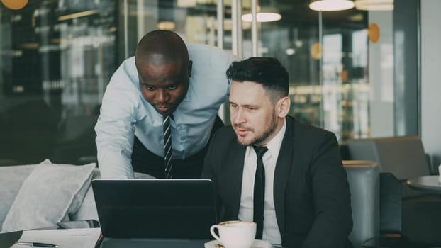 Two business partners discussing actively their startup project sitting in stylish cafe. Bearded Caucasian businessman sitting ang talking African American colleague while looking at laptop computer