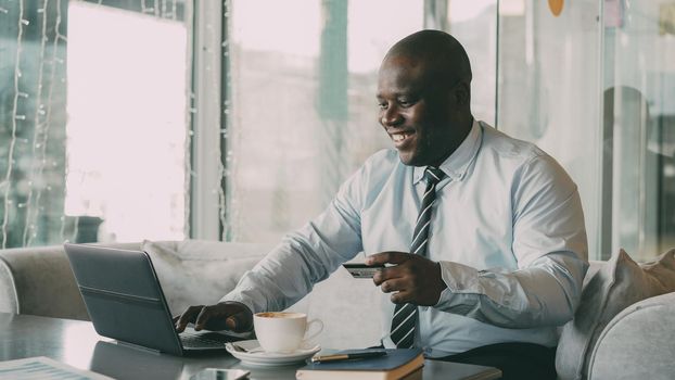 Optimistic African American businessman in formal clothes paying online bill using his credit card and laptop in fashionable cafe during lunch breaak. He smiles happily and raises his clenched fist.