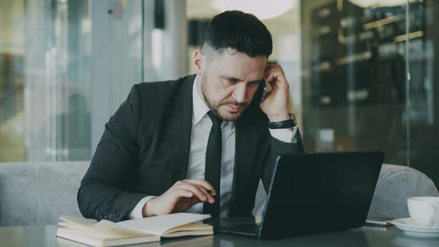 Smart Caucasian businessman sitting, talking on smartphone and writing down business information in his notepad while looking at laptop in glassy cafe