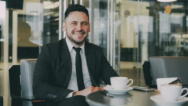 Portrait of cheery bearded Caucasian businessman in formal clothes sitting, drinking coffee and smiling sincerely in modern cafe having lunch break. Smartphone, pen and coffee cup sre on his table