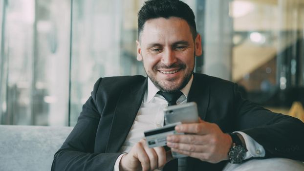 Cheerful Caucasian businessman in suit using online banking holding credit card and smartphone in his hands in modern cafe indoors during lunch break