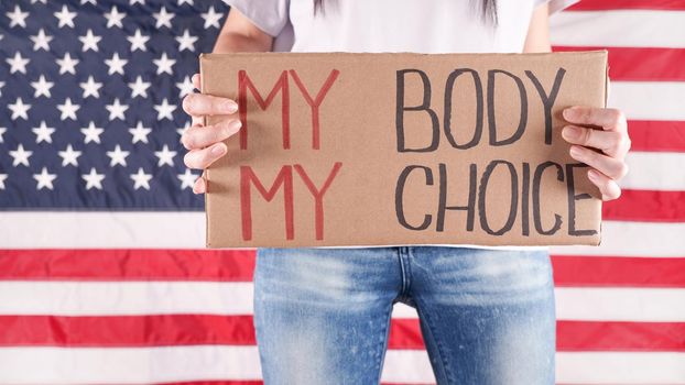 Young woman protester holds cardboard with My Body My Choice sign against USA flag on background. Girl protesting against anti-abortion laws. Feminist power. Equal opportunity Womens rights reedom