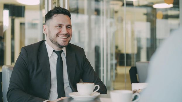 Optimistic Caucasian businessman smiling, gesticulating and rejoicing in buisness profits while speaking to his colleague in mod cafe during lunch. Coffee cups are on their table