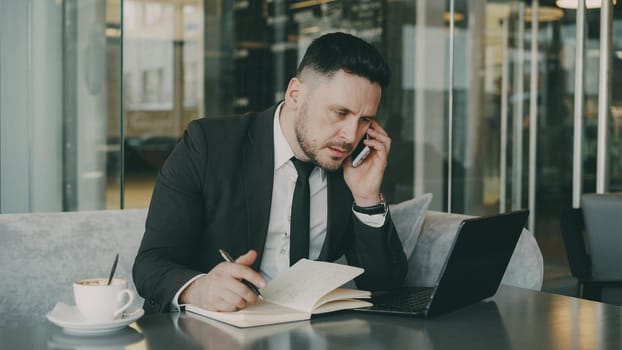 Bearded Caucasian businessman using laptop, talking on smartphone and writing down business information in modern cafe indoors during lunch break