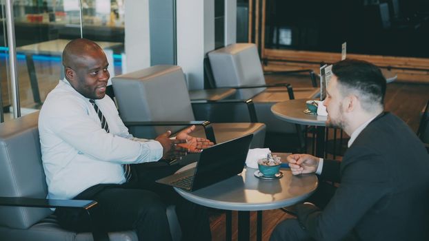 Two businessmen of Caucasian and African American in formal clothes smiling, gesturing and discussing their startup in spacious cafe during lunch break. Innovative laptop is on their table.