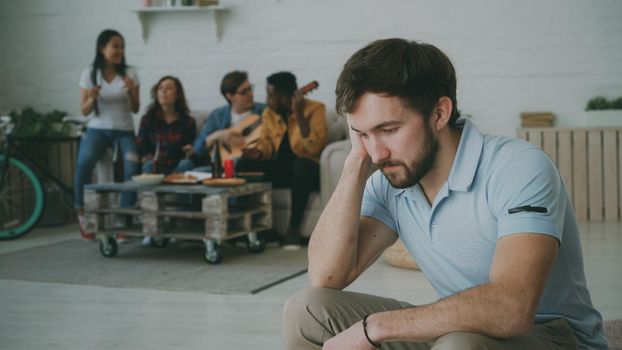 Young student guy feels upset and isolated while his friends celebrating party at home