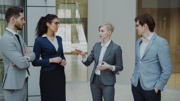 Businesswoman talking to her colleagues while standing in office lobby indoors. Group of business people discussing future deal