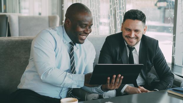 Cheerful African American businessman in formal clothes using digital tablet discussing startup project with his caucasian partner in stylish cafe indoors during lunch break