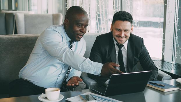 Cheerful African American businessman in formal clothes discussing business project with his caucasian colleague on his laptop in stylish cafe during lunch break