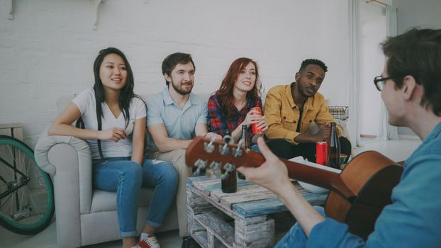 Happy young friends have party at shared apartment and singing together while their friend playing guitar at home indoors