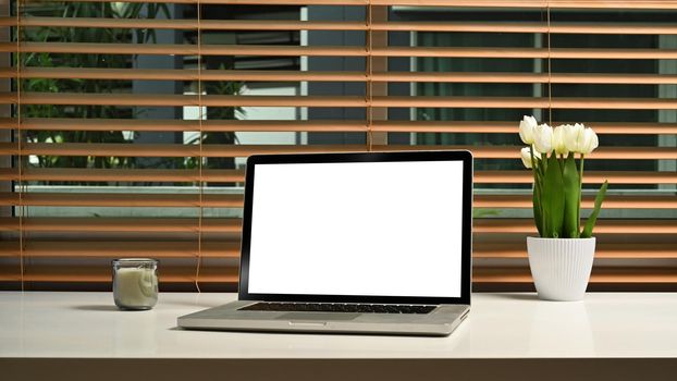 Home office desk. Computer laptop with blank screen, flower pot and candle on white table.