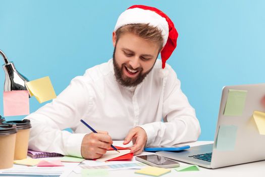 Smiling man in santa claus hat writing down letter or signing greeting postcard in red envelope, romance way to congratulate with holidays. Indoor studio shot isolated on blue background