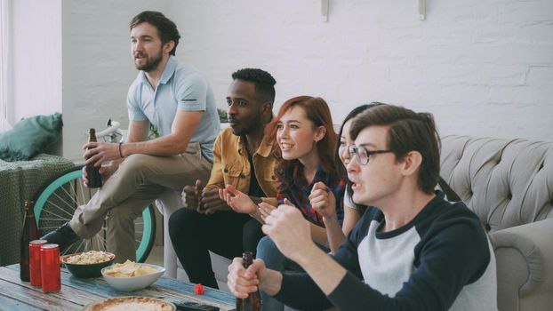 Multi ethnic group of friends sports fans watching sport event on TV together eating snacks and drinking beer at home indoors on holidays