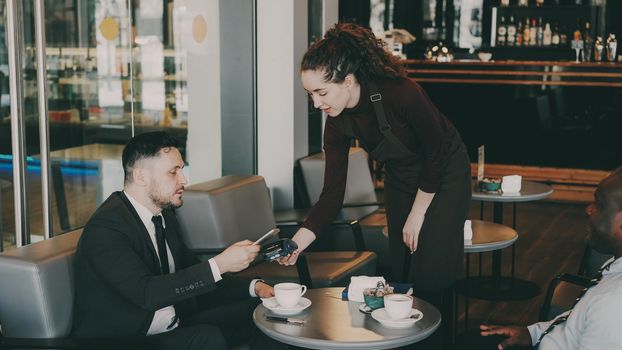 Happy Caucasian businessman smiling, sitting with his African American partner and paying online bill using his smartphone in modern cafe. Optimistic young waitress using her portable cash terminal.