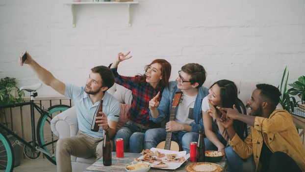 Happy student friends taking selfie and posing while have party with beer and pizza in shared accommodation