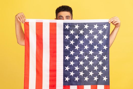 Man with big eyes peeking from big flag of united states of america he holding in hands, proud of his social security in country, feeling in safe. Indoor studio shot isolated on yellow background