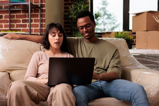 Diverse couple looking at decor inspiration on internet website, buying furniture to decorate new rented household. Shopping to move in apartment property and start beginnings.