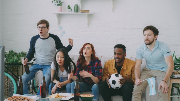 Multi-ethnic group of friends sports fans with Argentinian flags watching football championship on TV together at home indoors and cheering up favourite team