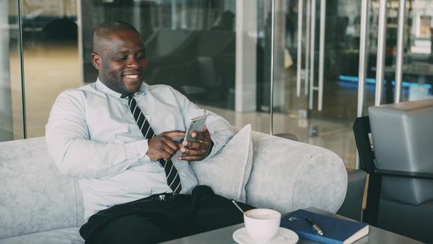 Cheerful African American businessman in formal clothes using smartphone in cafe. He looks at screen and smiles happily, with a cup of coffee on his table.