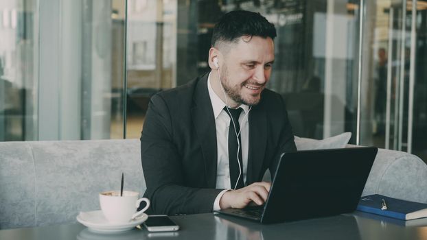 Portrait of cheery Caucasian businessman in formal clothes sitting and typing on his laptop while listening to music with earbuds in his ears in mod cafe. He smiles and shakes his head happily.