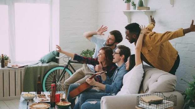 Multi ethnic group of cheerful friends taking selfie photos on smartphone camera while celebrating at party with beer and snacks at home