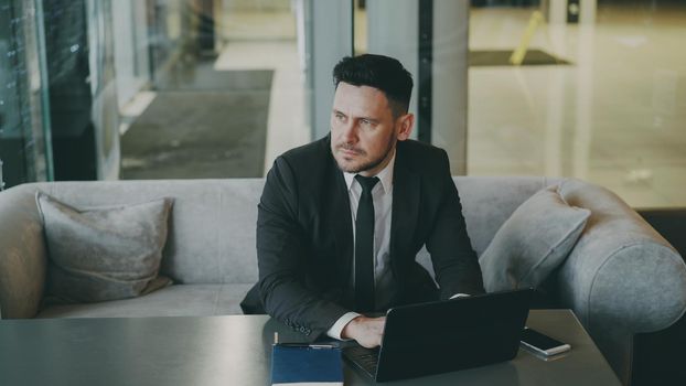 Upset thoughtful bearded Caucasian businessman sitting and thinking over his business plan while looking at his laptop screen in glassy cafe. Smartphone, notepad and coffee cup are on his table.