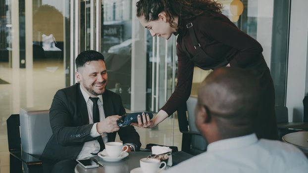Successful Caucasian businessman smiling, sitting with African American investor and paying online bill with his smartphone in modern cafe. Happy young waitress using her portable cash terminal.