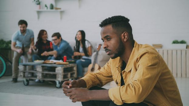 Young african amercan man feels upset and isolated while his friends celebrating party at home