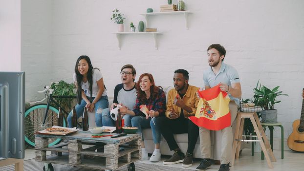 Multi-ethnic group of friends sports fans with Spainsh flags watching football championship on TV together at home indoors and cheering up favourite team
