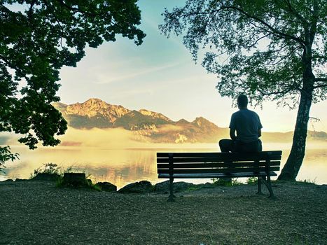 Alone man sits on bench beside an azure mountain lake. Man relax and watch high peaks of Alps above lake mirror.