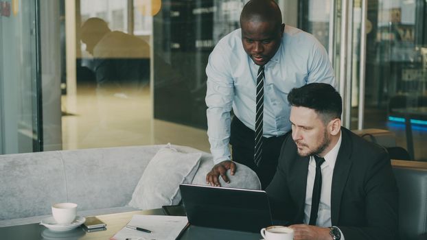 Two business partners discussing actively their startup project sitting in stylish cafe. Bearded Caucasian businessman sitting ang talking African American colleague while looking at laptop computer