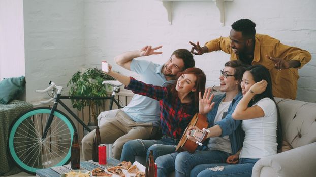 Group of joyful friends taking selfie photos on smartphone camera while celebrating at party with beer and snacks at home