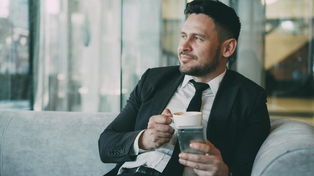 Smiling bearded Caucasian businessman in formal clothes smiling, relaxing and looking at his smartphone screen while drinking coffee in airy cafe during lunch time