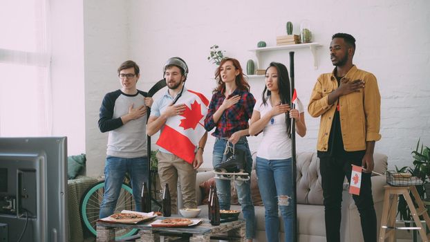 Multi ethnic group of friends sport fans listening and singing national Canadian anthem before watching sports championship on TV together at home indoors