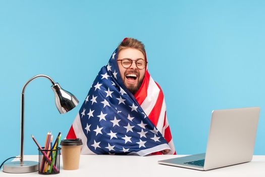 Extremely happy funny man employee sitting wrapped in American flag and shouting for joy in office workplace, celebrating labor day or US Independence day 4th of july, government employment support.