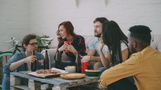 Multiracial young cheerful friends enjoying sitting together and talking each other while drinking beer and eating snacks at home indoors