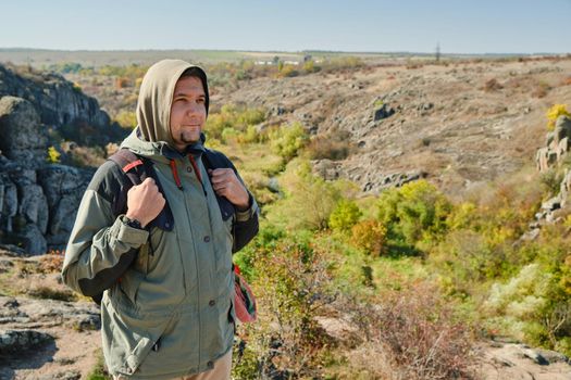 Hiker young man with backpack and trekking poles standing on edge of cliff and looking at the mountains in summer outdoor