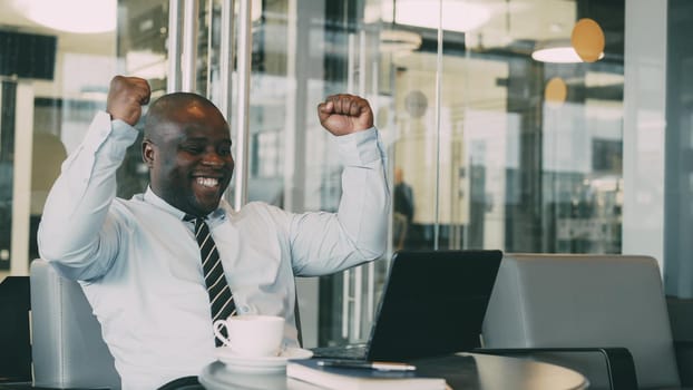 Successful african american businessman using laptop computer receiving good message and become very excited and dancing while sitting in modern cafe duiring coffee braek