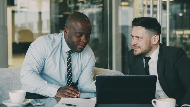 Two business colleagues looking at laptop computer and discussing their startup plan in modern office with glass walls. Bearded businessman and his partner sitting at table and talking future deals