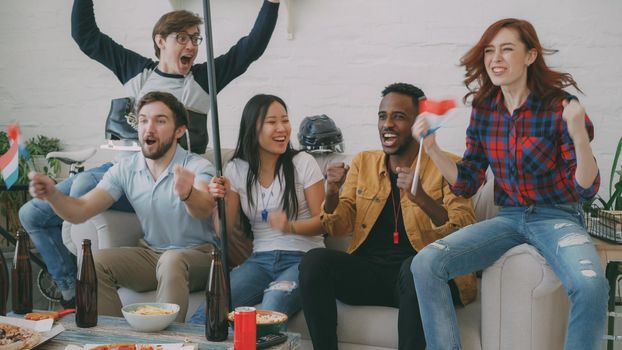 Diverse group of friends sport fans with Dutch national flags watching winter sports games on TV together cheering up favourite team at home indoors