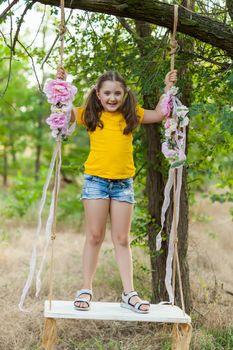 Cute smiling girl in a yellow t-shirt having fun on a swing in tree forest. Sunny day. Summer outdoor activities for kids