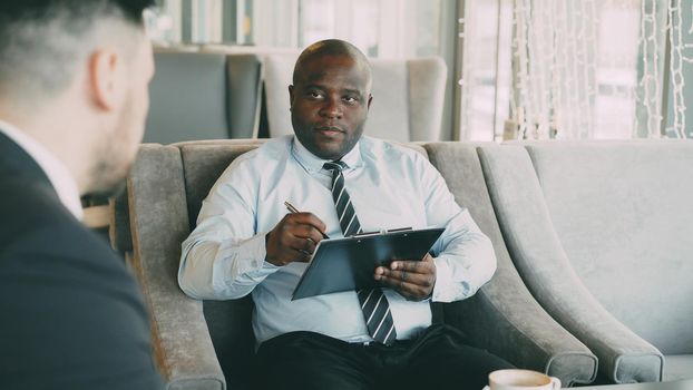 African american HR manager having job interview with young man in suit and watching his resume application in modern cafe indoors