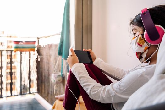 little girl sitting in front of her home window with a headset and protective mask watching videos on the phone, coronavirus concept and home entertainment for children, copy space for text