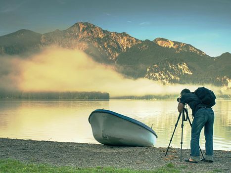 Man hiker is taking photo of ship at mountain lake shore. Silhouette at fishing paddle boat at lake coast.