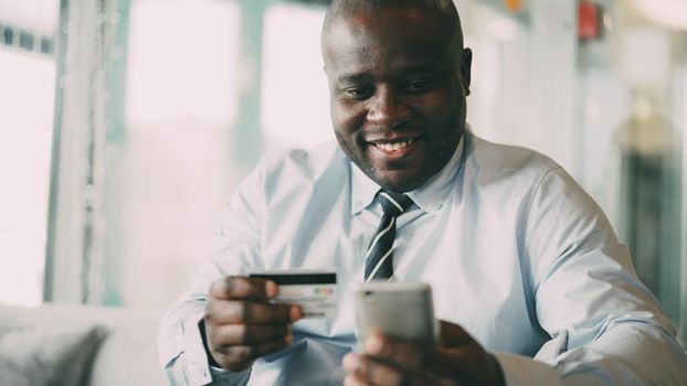 Happy African American businessman in formal clothes smiling and paying online bill while keeping credit card and smartphone in his hands in glassy cafe